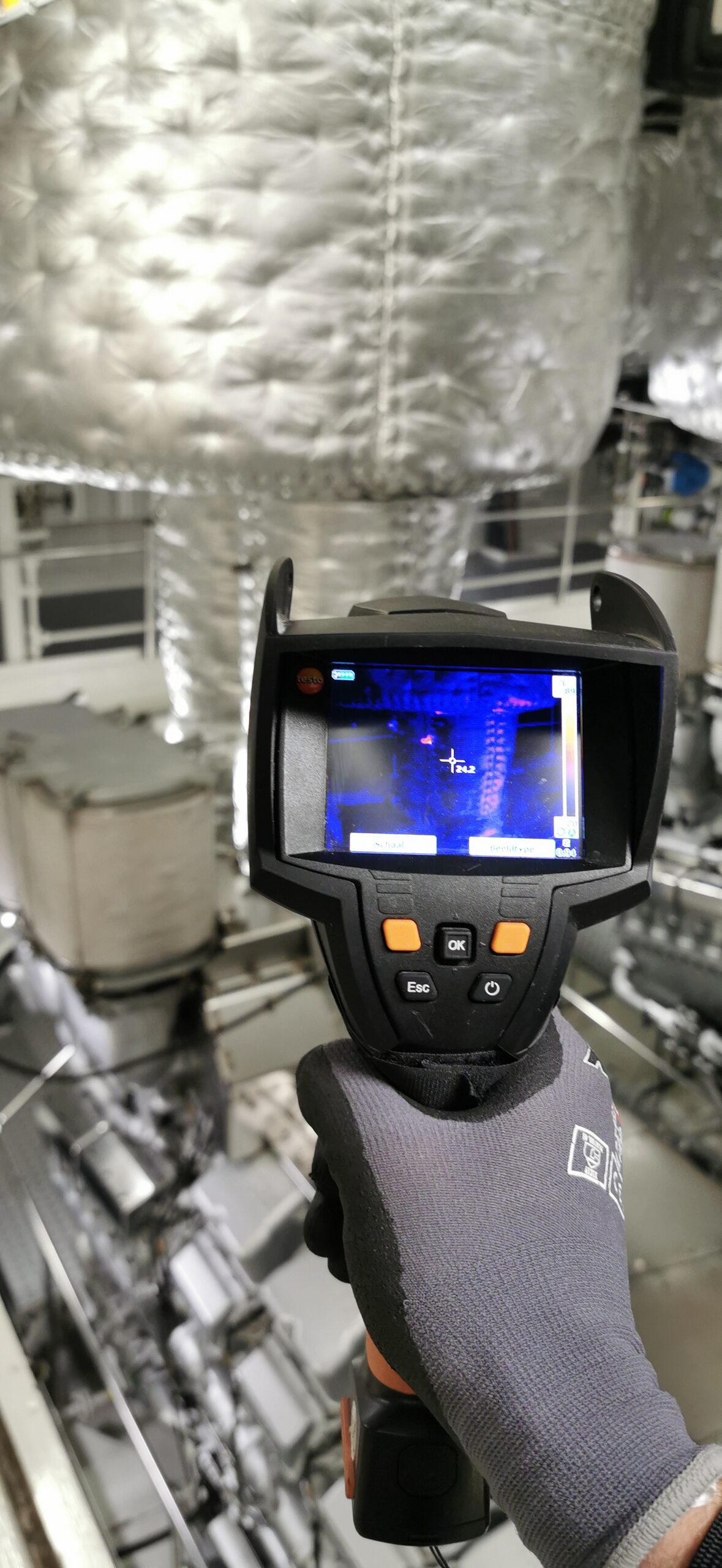 Hand in werkhandschoen houdt een warmtebeeldcamera onder geïsoleerde uitlaatleidingen in een machinekamer
