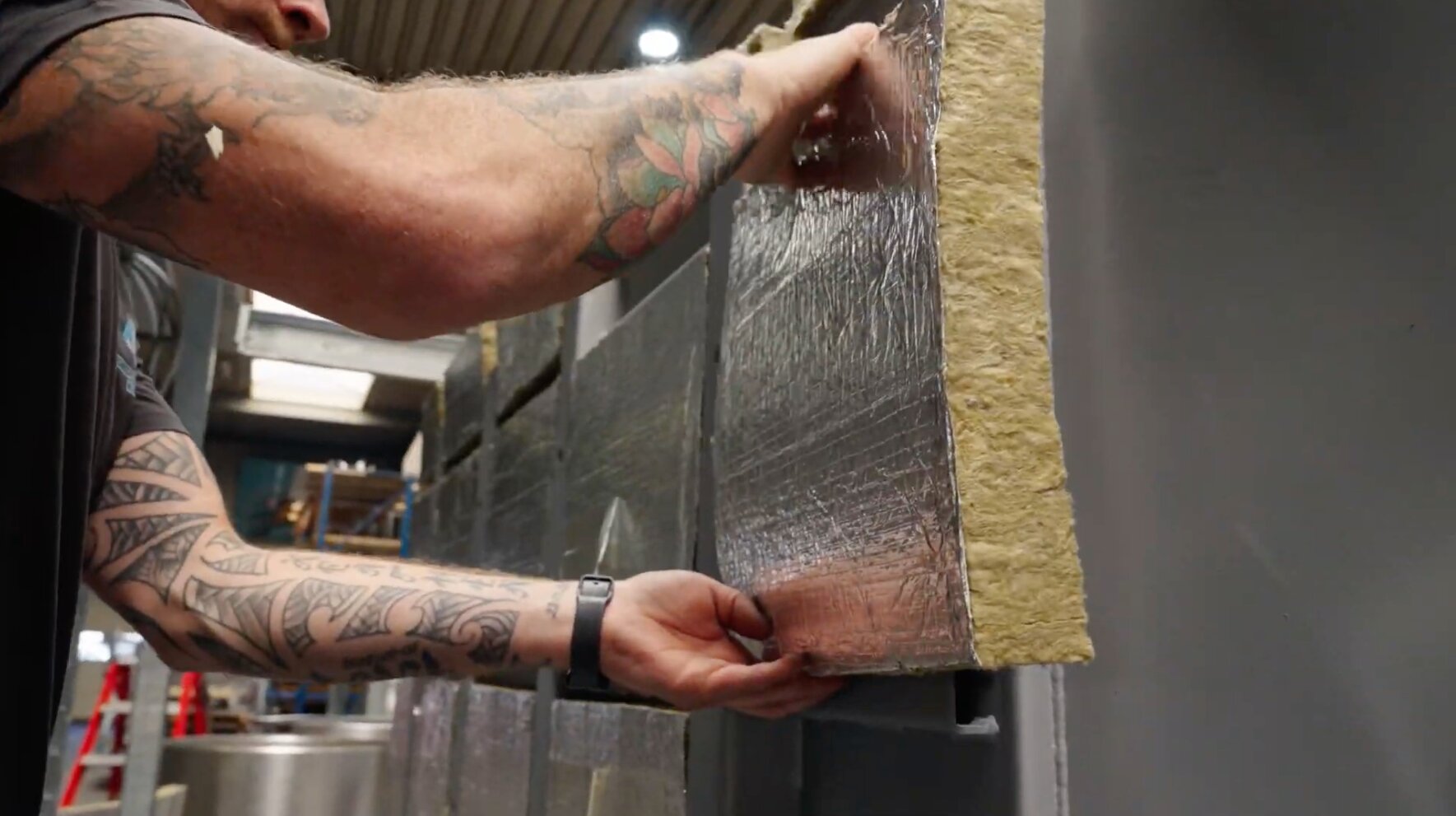 Medewerker brengt steenwolplaten met aluminiumfolie aan op een stalen wand in de werkplaats